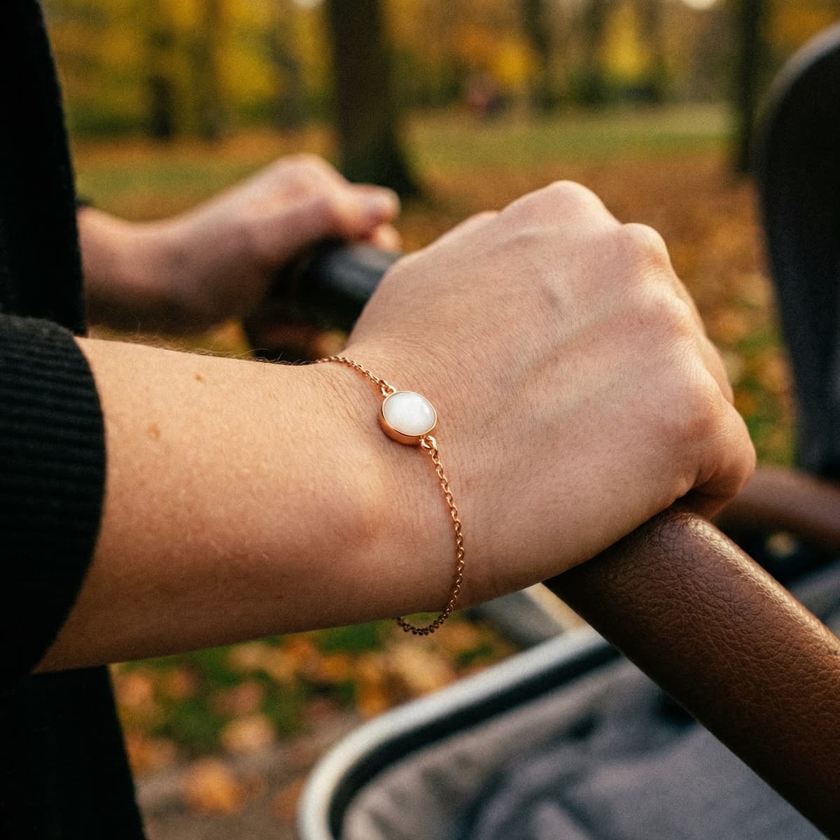 Zartes roségoldenes Armband mit Muttermilch und weißem rundem Stein am Handgelenk, im Hintergrund ein unscharfer Kinderwagen in einem herbstlichen Park.