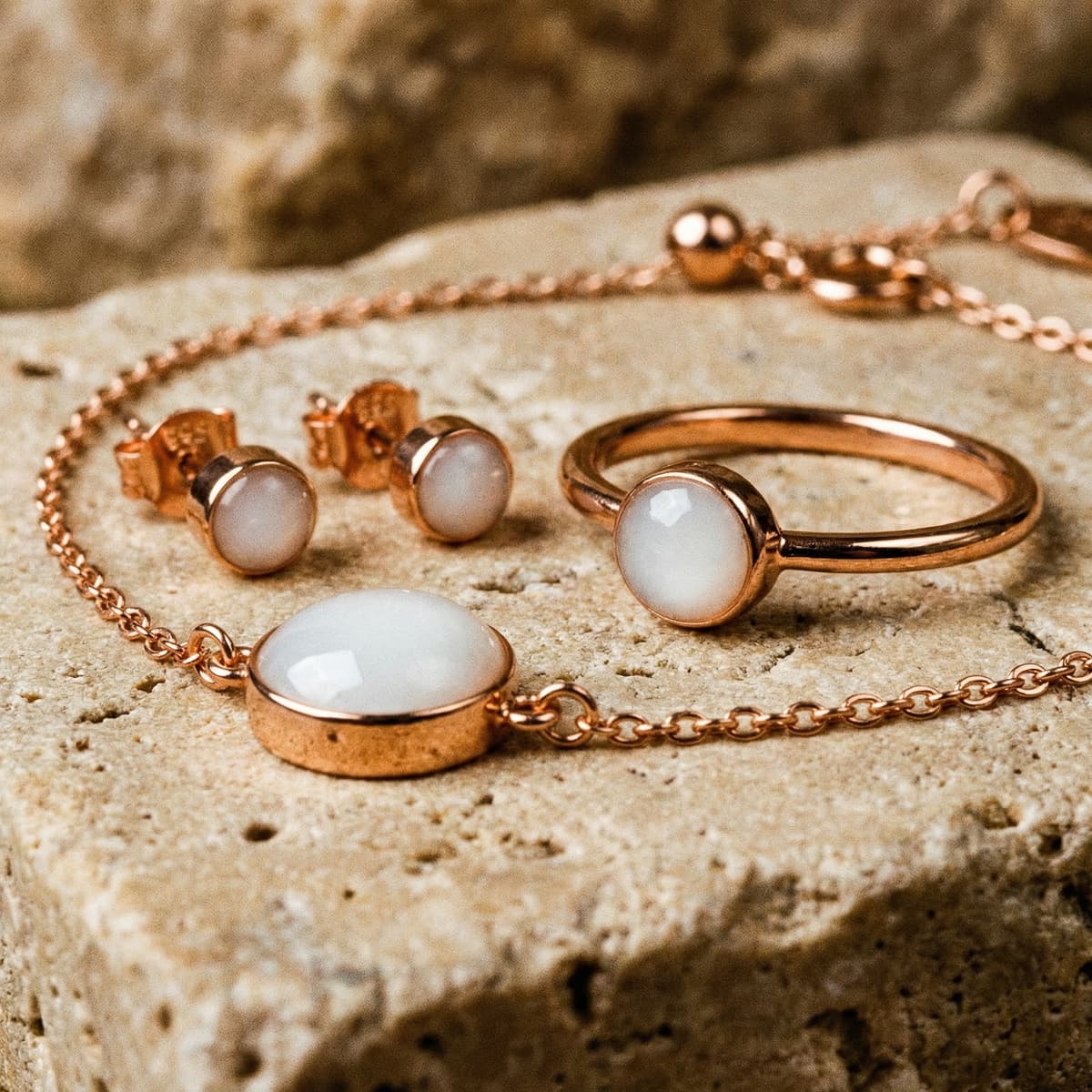 Roségoldenes Schmuckset mit Armband Muttermilch, passendem Ring und Ohrsteckern mit milchig-weißem Harzstein, arrangiert auf natürlichem Steinuntergrund.