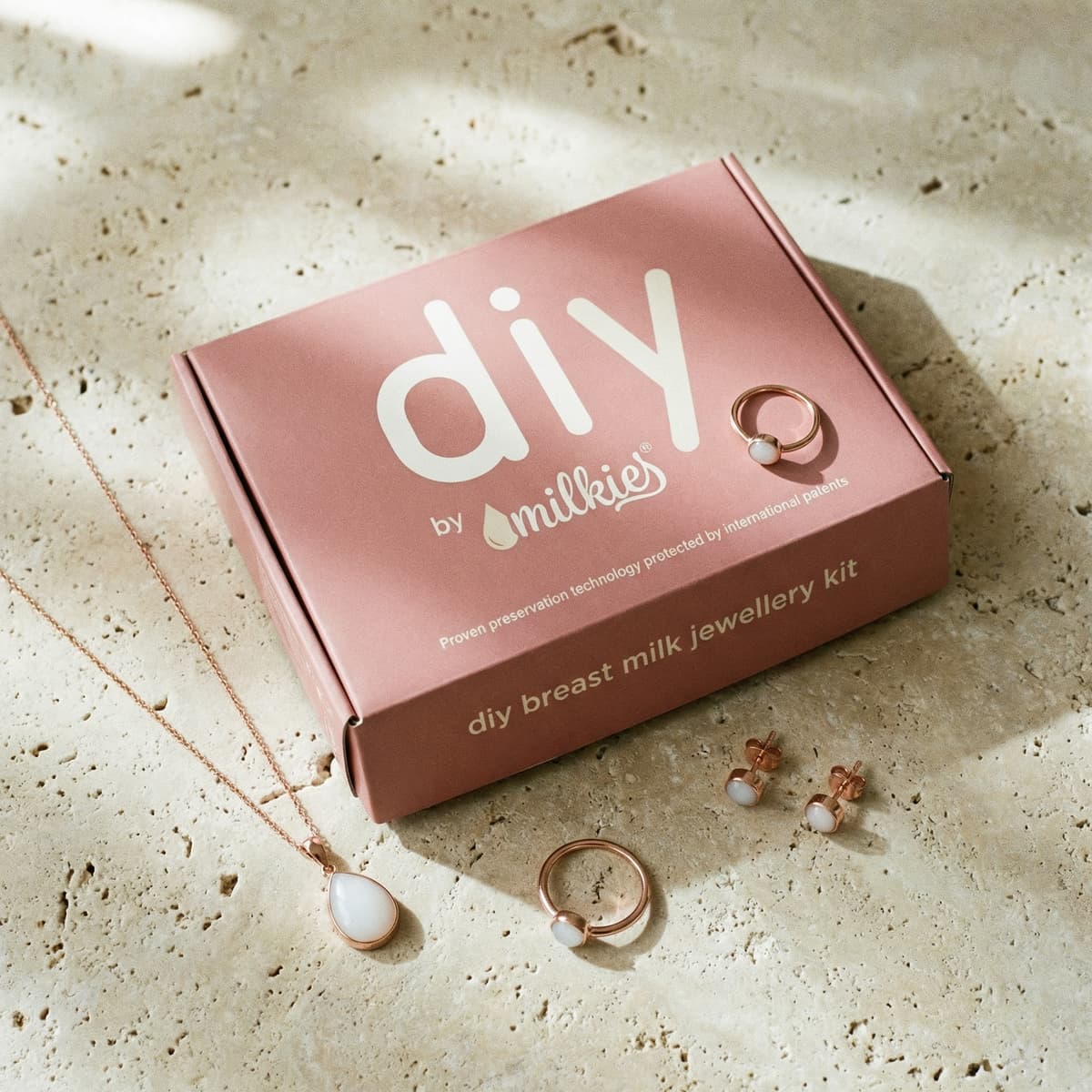 Rosafarbene Box mit DIY by MILKIES DIY Resin Set für Muttermilch auf hellem Steinuntergrund, umgeben von Kette, Ringen und Ohrsteckern mit milchig-weißem Harzstein.