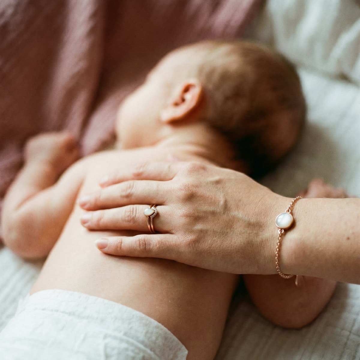 Nahaufnahme eines schlafenden Babys, während die Mutter schützend ihre Hand auf seinen Rücken legt und ein zartes Armband sowie einen Ring mit milchig-weißem Stein als Erinnerung an die Stillzeit trägt.
