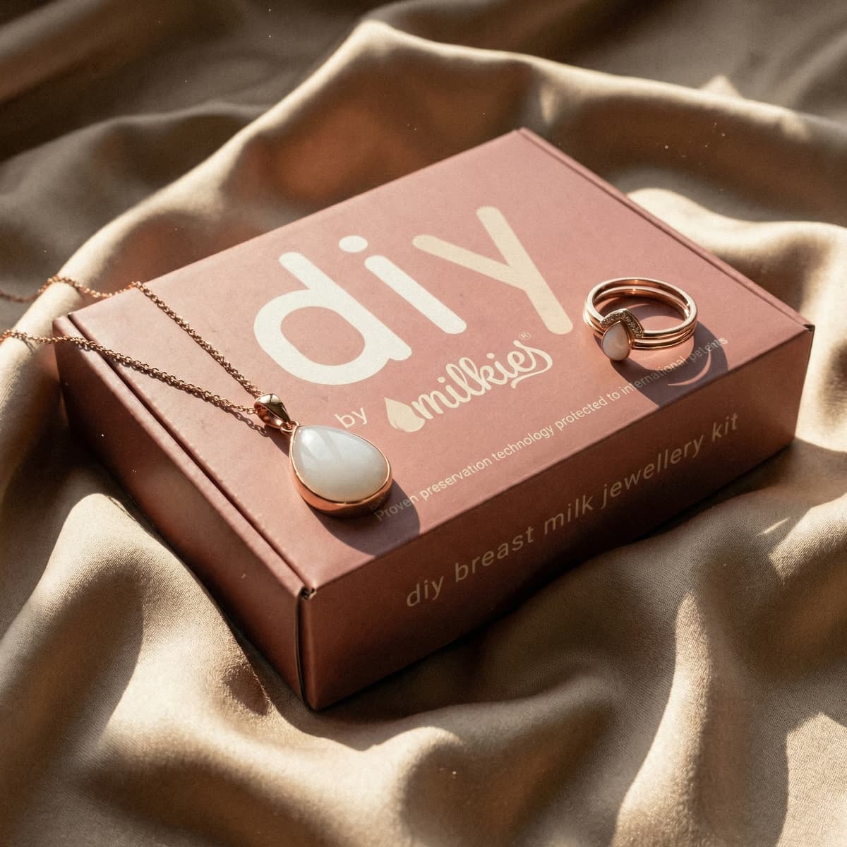 DIY by MILKIES Brustmilch-Schmuckset als geschenk für neue mutter – rosa Kit-Box auf weichem Stoff, daneben roségoldene Kette mit milchigem Anhänger und passender Ring
