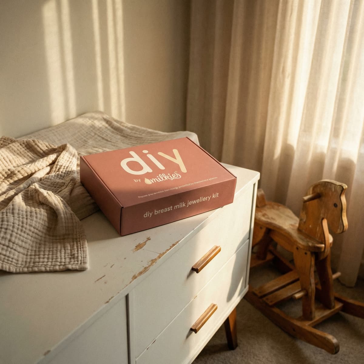 Stilvolles DIY by MILKIES Breastmilk Jewellery Kit in roséfarbener Geschenkbox – ein elegantes Geschenk für Schwangere – auf weißer Kommode im warm beleuchteten Schlafzimmer neben Decke und Holz-Schaukelpferd.