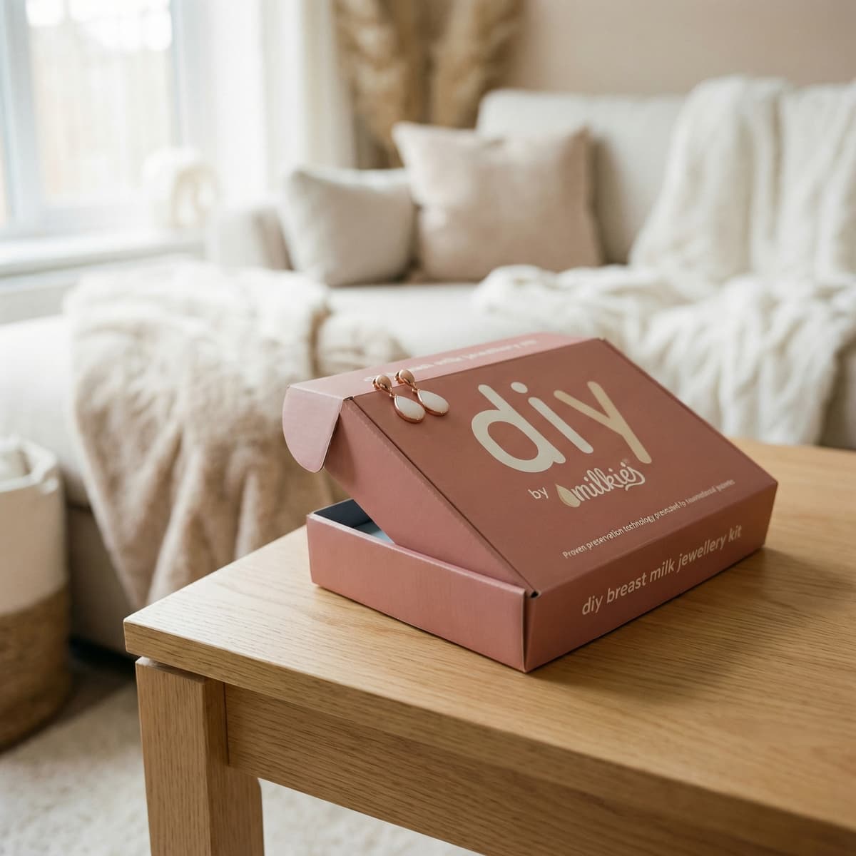 DIY by MILKIES Brustmilch-Schmuckkit in roséfarbener Box als stilvolles Geschenk zum Abstillen, auf einem Holztisch im gemütlichen Wohnzimmer mit Ohrringen obenauf.
