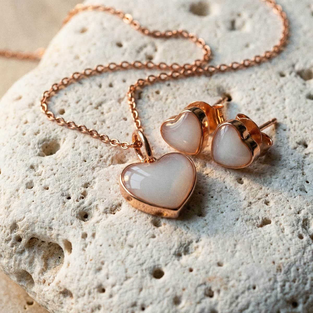 Herzförmiges Schmuckset in Roségold mit milchig-weißem Harzanhänger an Kette und passenden Ohrsteckern auf hellem Stein, elegantes geschenk zur geburt frau.