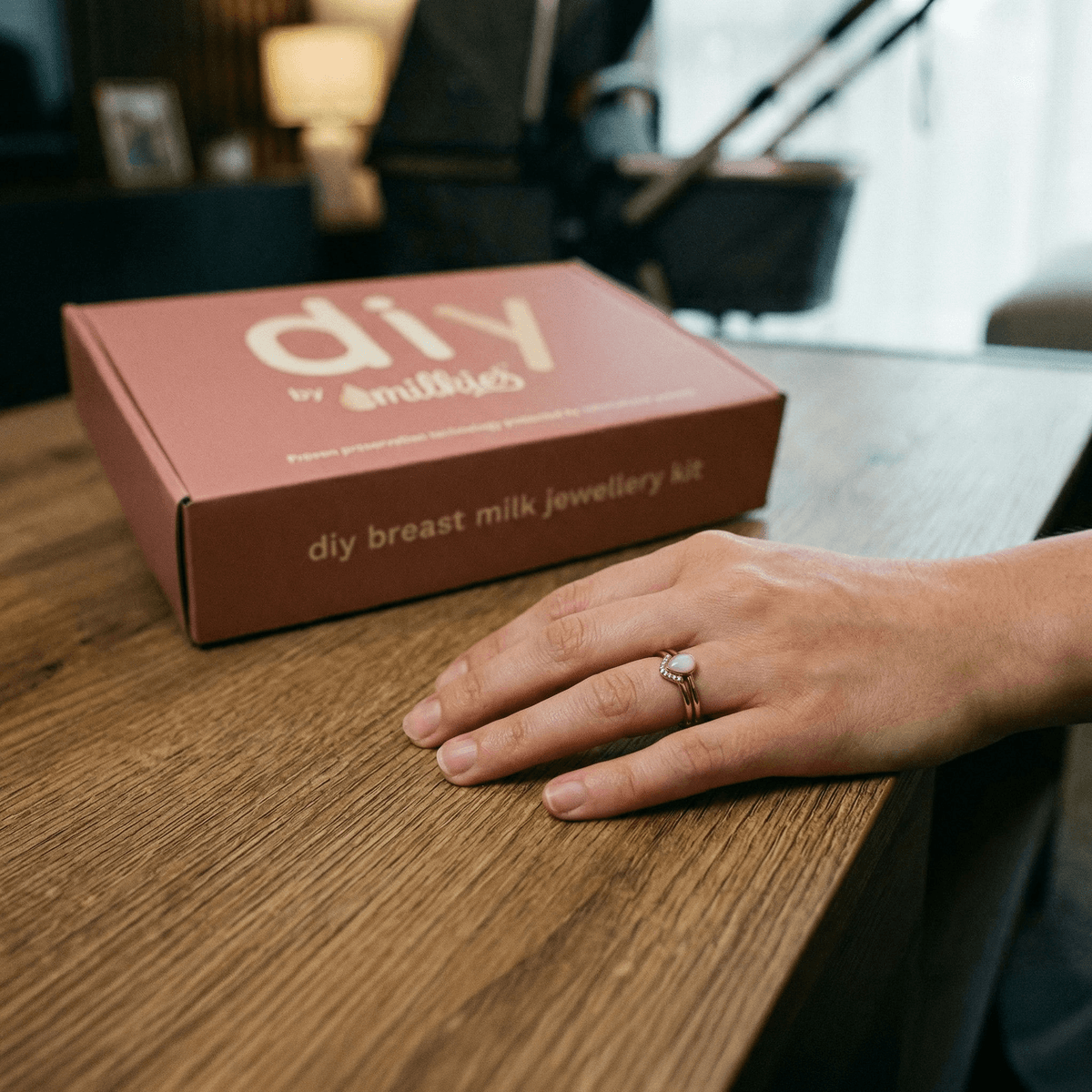 Hand mit Ring neben der DIY by MILKIES Breastmilk Jewellery Kit-Box auf Holztisch, persönliches geschenk zur geburt frau als Erinnerungsstück zur Stillzeit