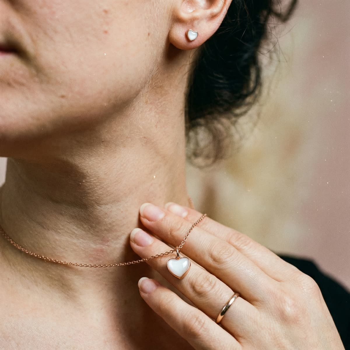 Frau trägt als Geschenk zur Stillreise eine zarte Kette mit herzförmigem, milchig-weißem Anhänger und passenden Ohrsteckern in Roségold-Optik.