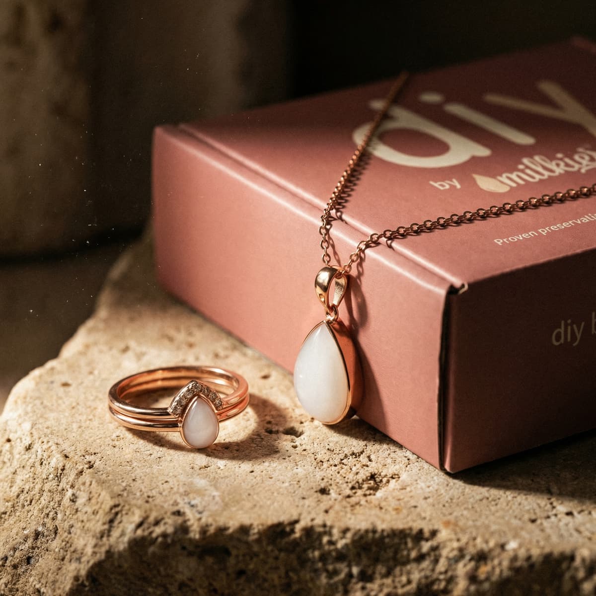Roségoldenes Schmuckset mit Ring und Tropfen-Halskette mit milchig-weißem Stein vor rosa DIY by MILKIES Box – persönliches Geschenk zur Stillreise.