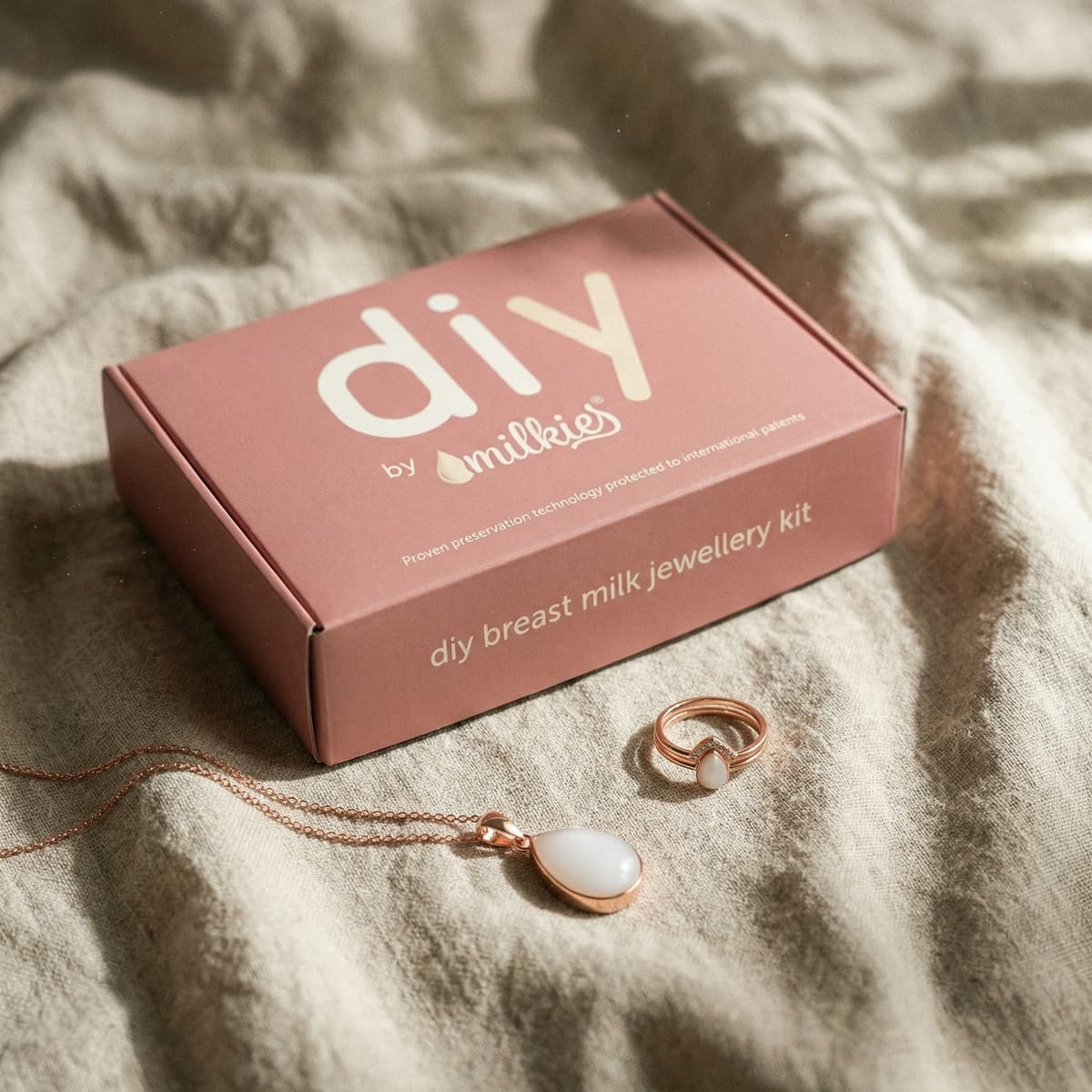DIY by MILKIES Brustmilch-Schmuckset als elegante Ideen für Geschenke zur Geburt: rosafarbene Box mit Kette und Ring in Roségold, auf weichem Stoff arrangiert.