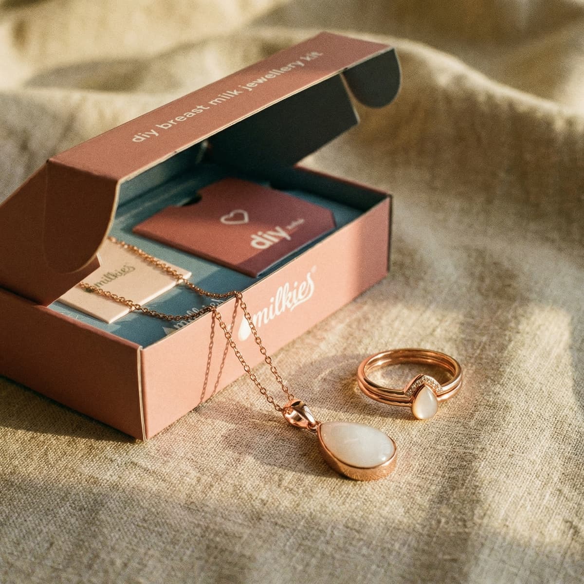 Roségoldenes Brustmilch-Schmuckset von DIY by MILKIES als mutter geschenk zur geburt: geöffnete Box mit Kette und Ring mit milchig-weißem Tropfenstein auf beigem Stoff