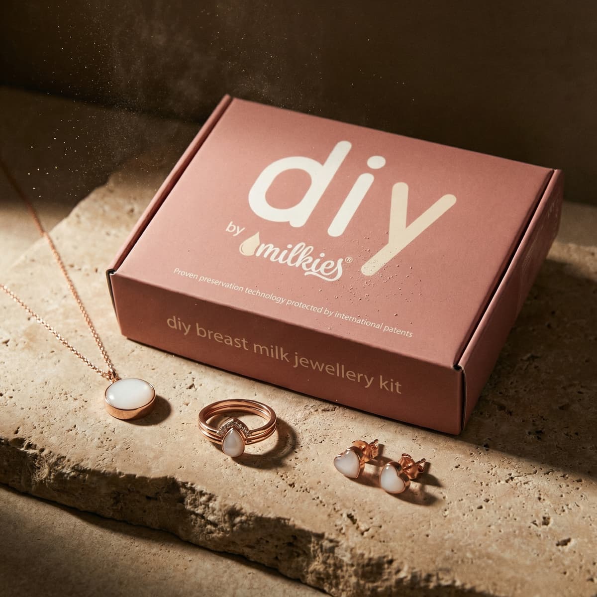 Stilvolle Produktaufnahme eines DIY by MILKIES Kits in roséfarbener Box mit Schmuckbeispielen – ein Muttermilch Schmuck DIY Set mit Kette, Ring und Herz-Ohrringen in Roségold mit milchig-weißen Einsätzen.