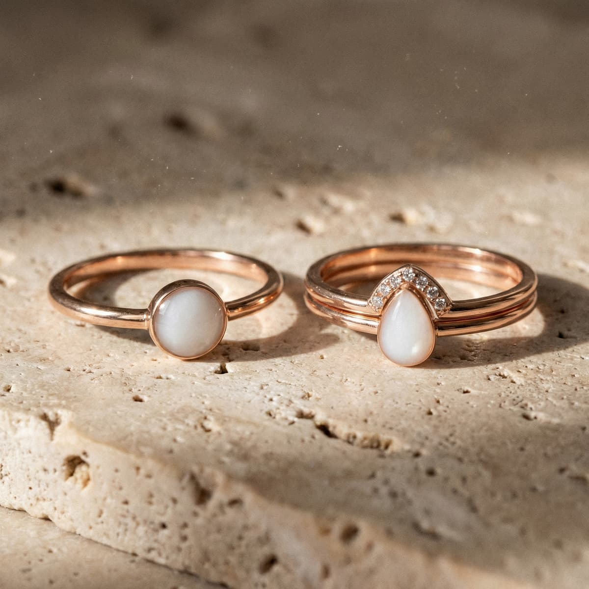 Roségoldene Ringe mit milchig-weißem Stein und kleinen Glitzersteinen, als muttermilch schmuck ring auf hellem Naturstein im warmen Licht fotografiert, einmal rund und einmal tropfenförmig.