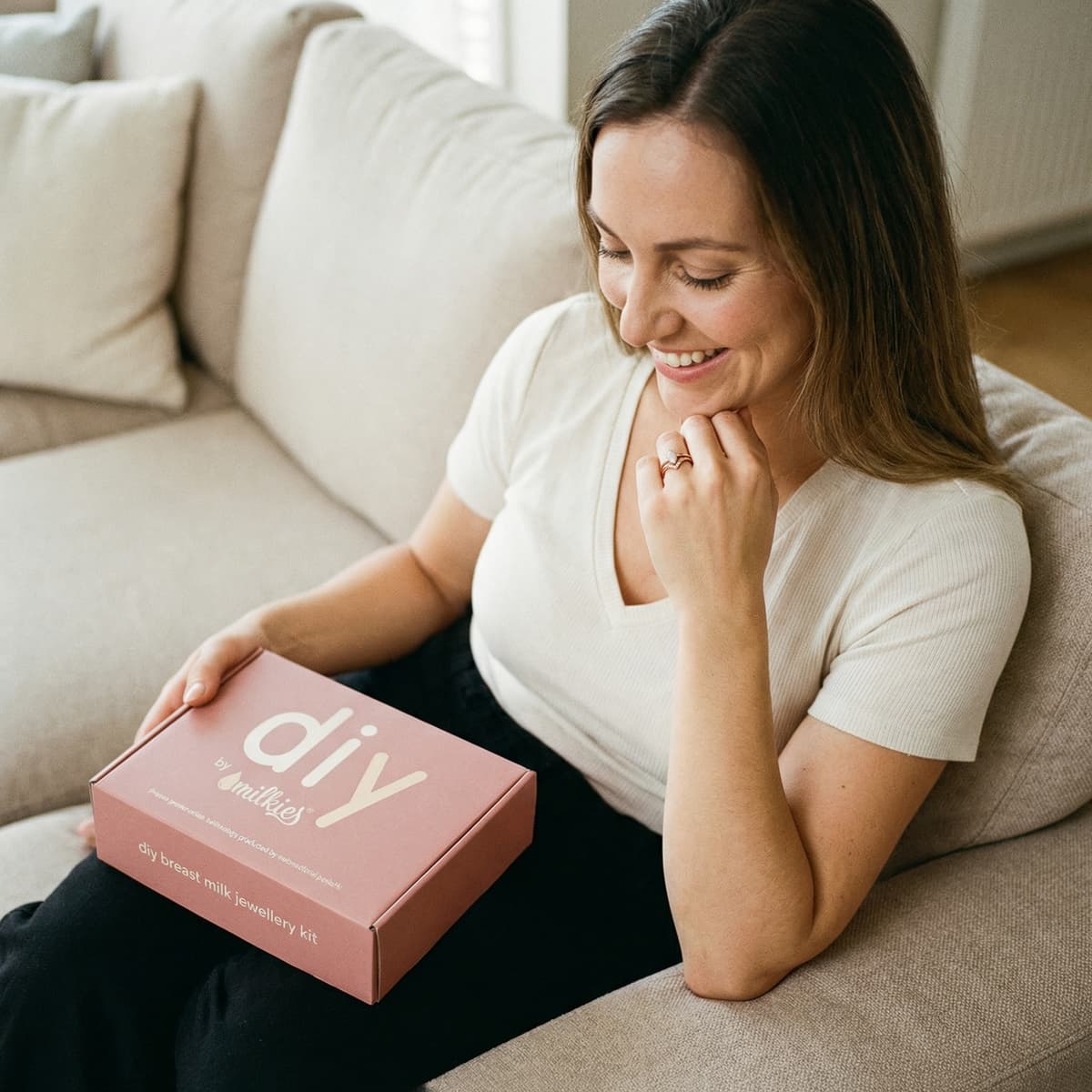 Glückliche Mutter auf dem Sofa mit DIY by MILKIES Schmuck-Kit in der Hand, ideal um einen muttermilch schmuck ring als persönliches Erinnerungsstück bequem und privat zu Hause herzustellen.