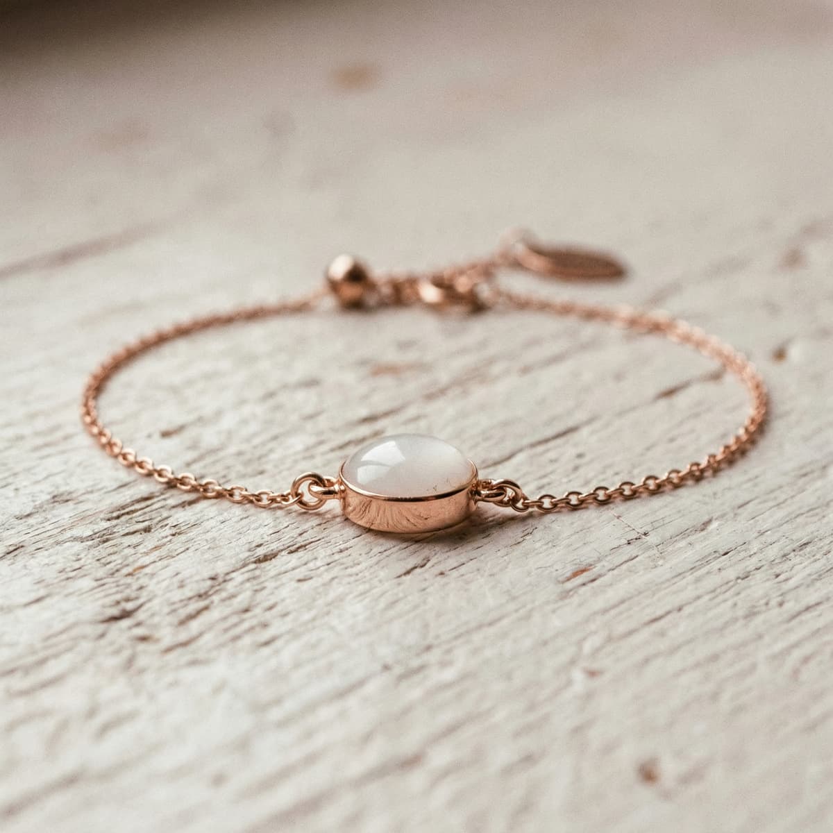 Zartes Armband in Roségold-Optik mit ovalem, milchig-weißem Cabochon-Anhänger auf Holzoberfläche – Beispiel für ein Bastelset für Muttermilch Armband als persönliches Erinnerungsstück.