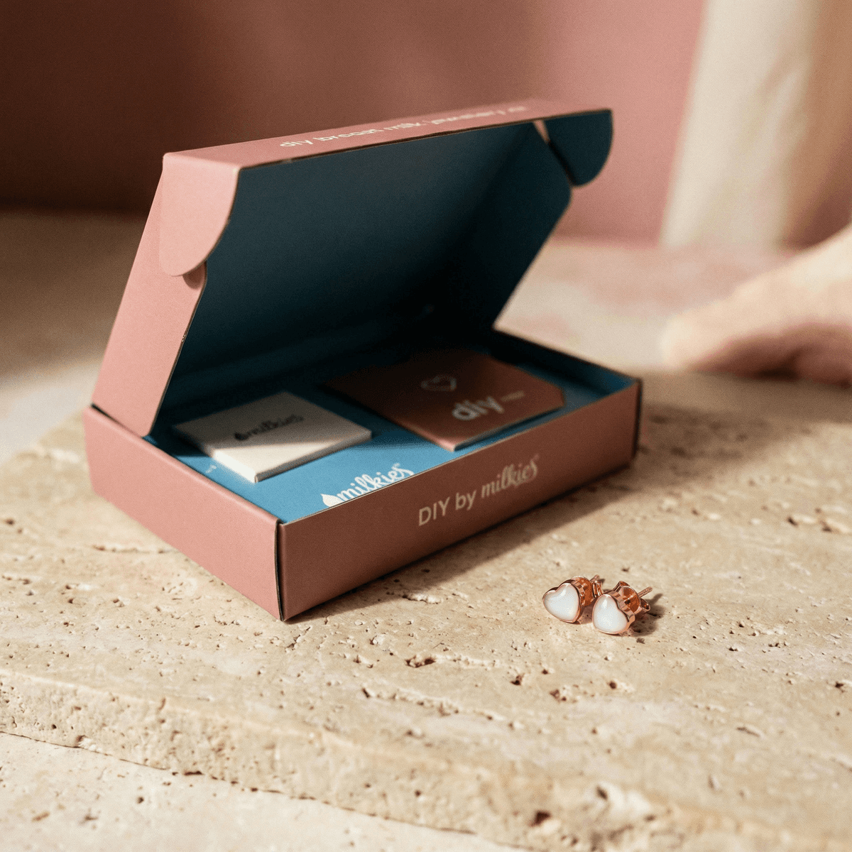 Geschenkbox von DIY by MILKIES mit Zubehör auf einer Steinoberfläche, daneben herzförmige roségoldfarbene Ohrstecker aus einem DIY Kit Muttermilch Ohrringe als personalisierbares Keepsake.