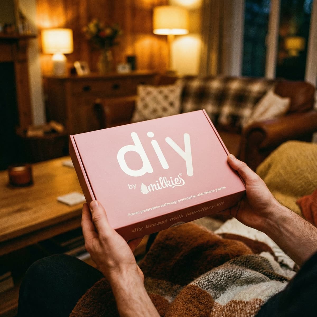 Hände halten eine rosafarbene DIY by MILKIES Box für ein Breastmilk-Schmuck-Set im gemütlichen Wohnzimmer – persönliches Geschenk für Ehefrau zur Geburt.