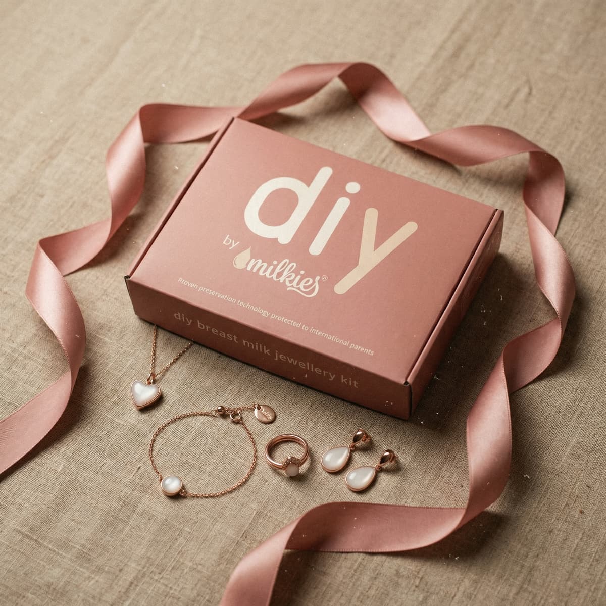 Rosa Geschenkbox mit Satinband und DIY by MILKIES Brustmilch-Schmuckset als Geschenk für frischgebackene Mama, mit Herz-Anhänger, Armband, Ring und Tropfen-Ohrringen.