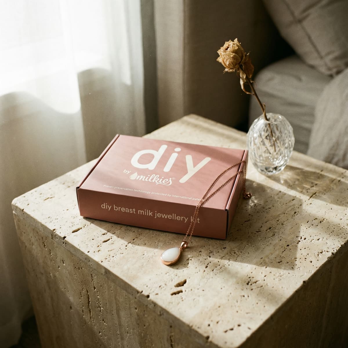 Rosafarbene Box mit DIY by MILKIES Brustmilch-Schmuckset als Geschenk zur Geburt, mit Kette und Anhänger auf einem Steintisch neben einer Glasvase mit getrockneter Rose.