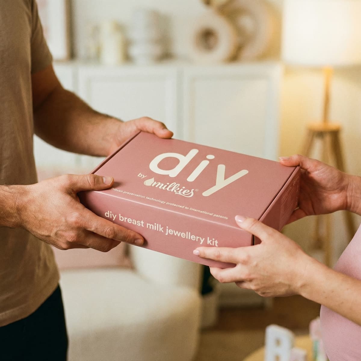 Ein Mann überreicht seiner Frau in einem warmen Wohnambiente ein geschenk zur geburt vom ehemann: die rosafarbene DIY by MILKIES Box „DIY Breast Milk Jewellery Kit“ für ein selbstgemachtes Muttermilch-Schmuckstück.