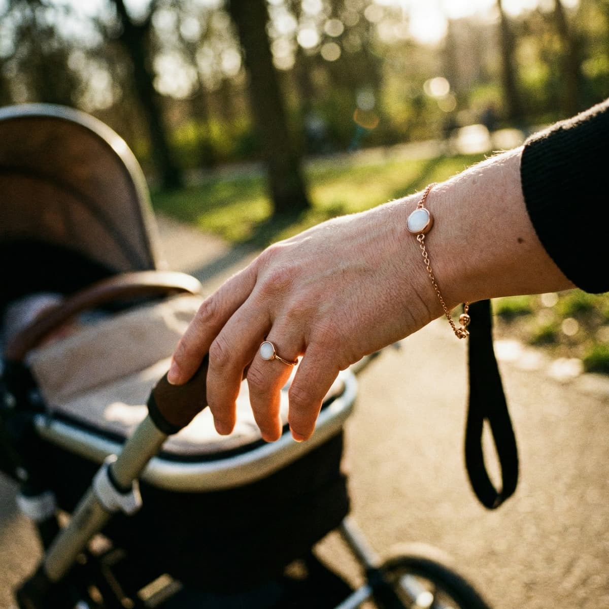 Hand einer Mutter am Kinderwagen im warmen Sonnenlicht mit zartem Armband und Ring als Muttermilch Andenken in roségoldener Fassung.