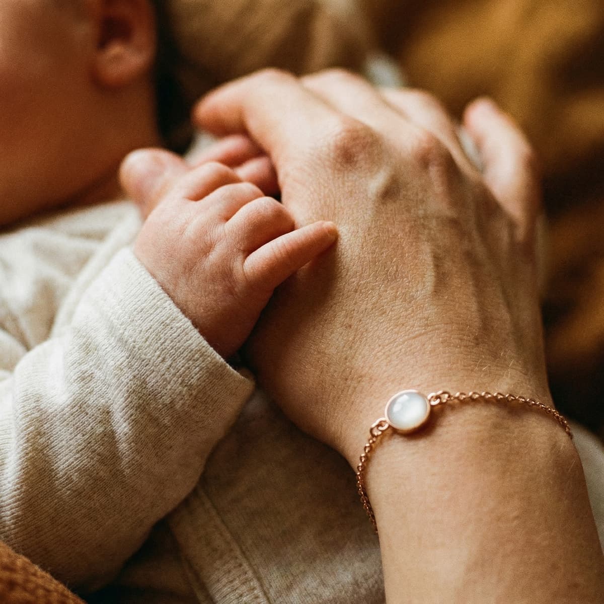 Nahaufnahme einer Mutterhand, die von einem Baby gehalten wird, mit einem zarten roségoldenen Muttermilch Armband und milchig-weißem Schmuckstein am Handgelenk.