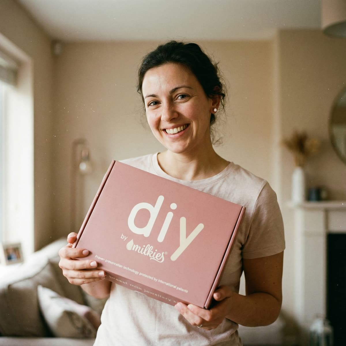 Lächelnde Mutter hält eine rosa DIY by MILKIES Box in der Hand, ein Muttermilch Ohrringe DIY Set, das zeigt, warum das Kit ideal ist, um zu Hause ein persönliches Erinnerungsstück selbst zu gestalten.