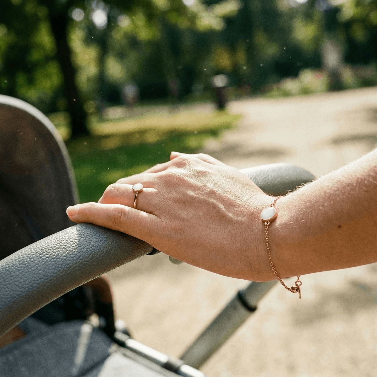 Hand einer Mutter am Kinderwagen im Park, trägt zarten Muttermilchschmuck: goldfarbenes Armband und Ring mit milchig-weißem Stein im Sonnenlicht