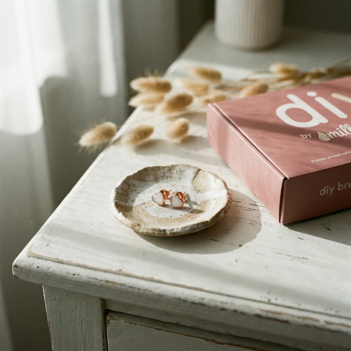 Roségoldene Ohrstecker mit milchig-weißen Steinen in einer Keramikschale neben einer rosa DIY by MILKIES® Box auf hellem Holztisch – Inspiration, wie man Ohrringe aus Muttermilch selber machen kann.