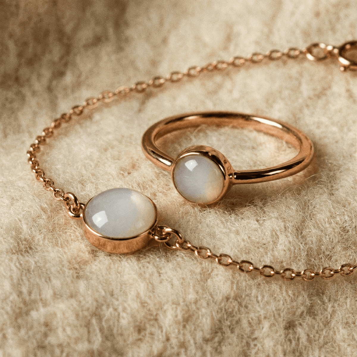 Roségoldfarbener Ring und filigranes Armband mit milchig-weißen Cabochons, darunter ein Ring aus Muttermilch, auf cremefarbenem Stoff im Close-up.