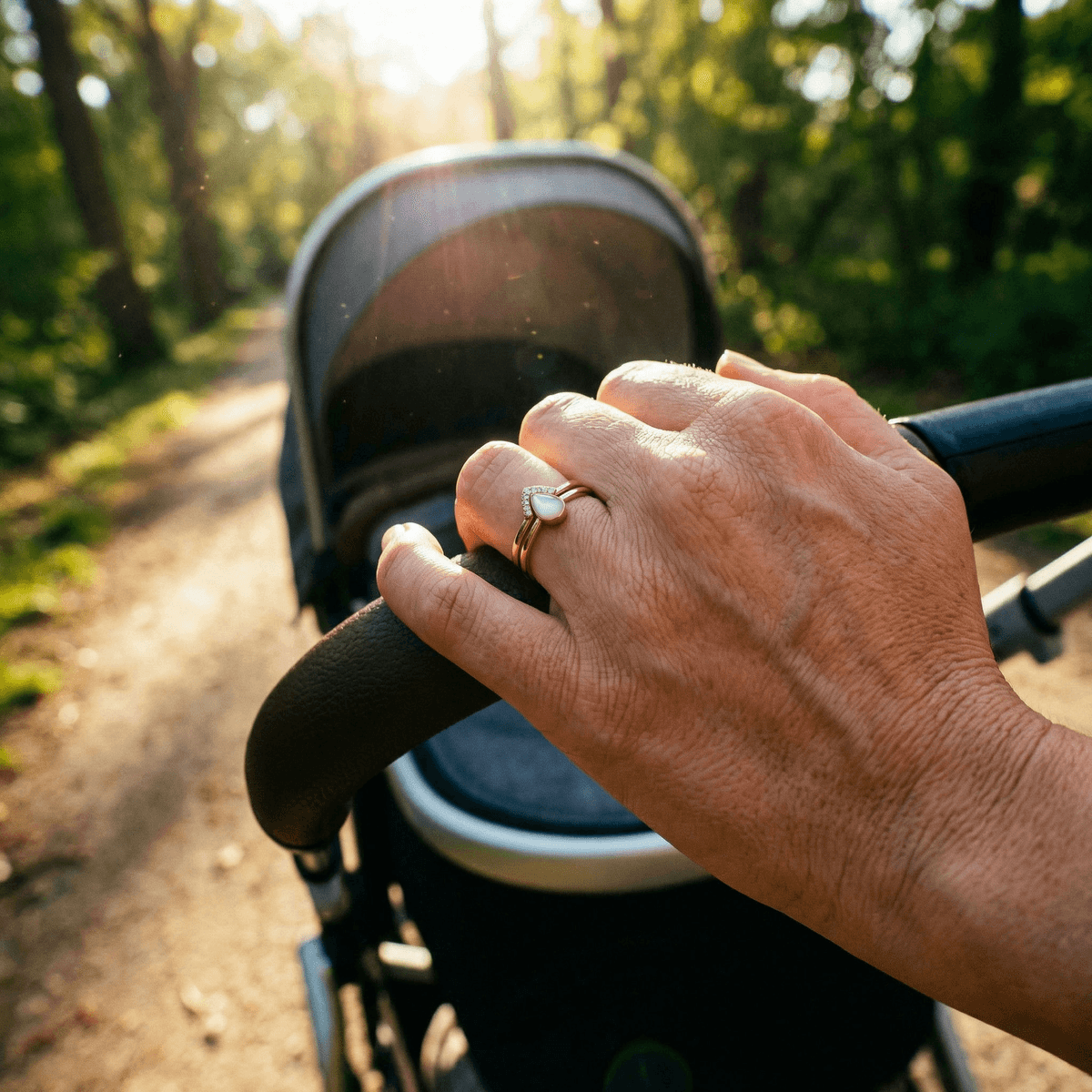 Hand mit zartem goldfarbenem Ring mit milchig-weißem Stein, ein Ring aus Muttermilch, am Kinderwagen bei einem Spaziergang im Park im warmen Sonnenlicht.