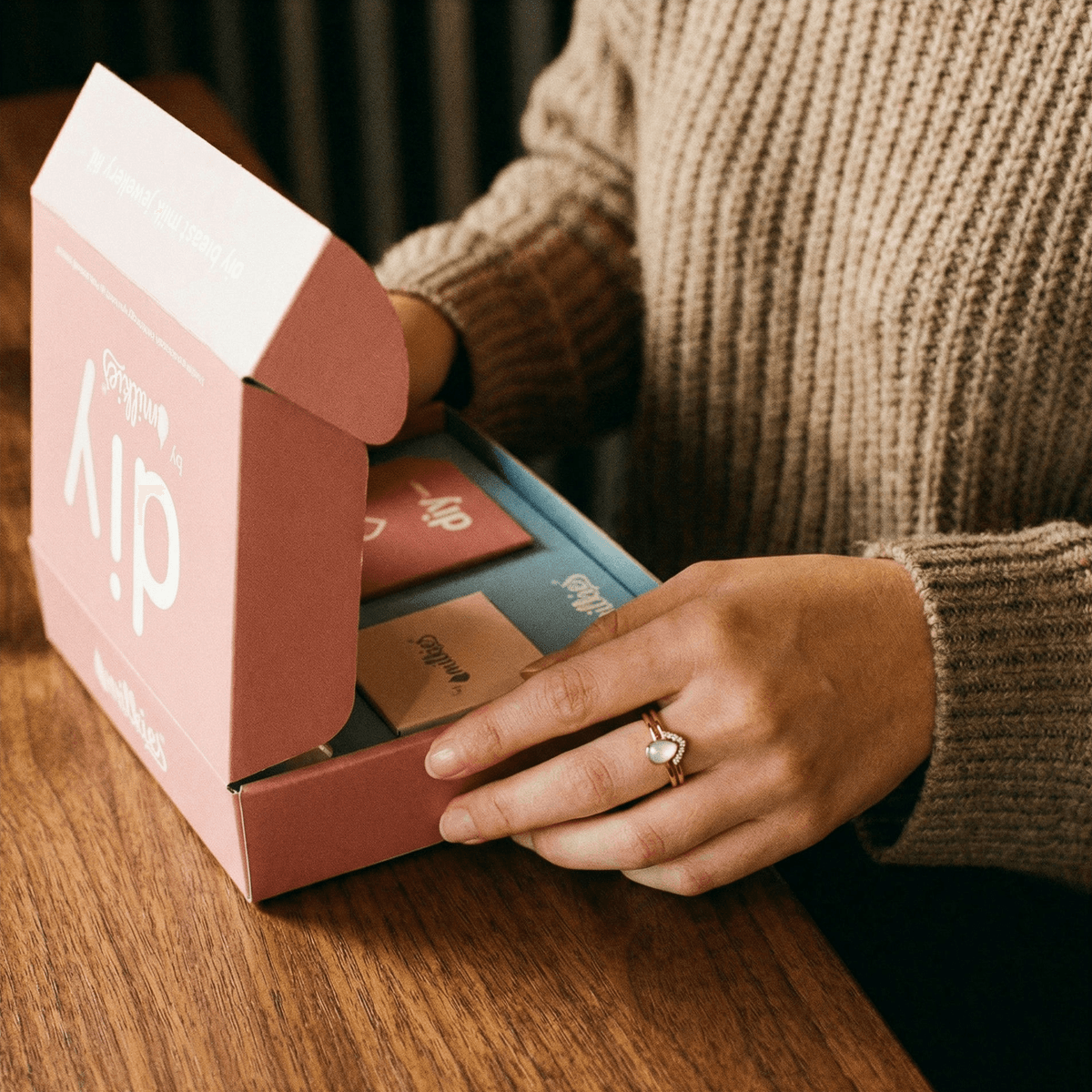 Hände öffnen das DIY by MILKIES Set auf einem Holztisch – ideal, um zuhause einen Ring aus Muttermilch als persönliches Andenken selbst zu gestalten und von dem praktischen DIY-Kit zu profitieren.